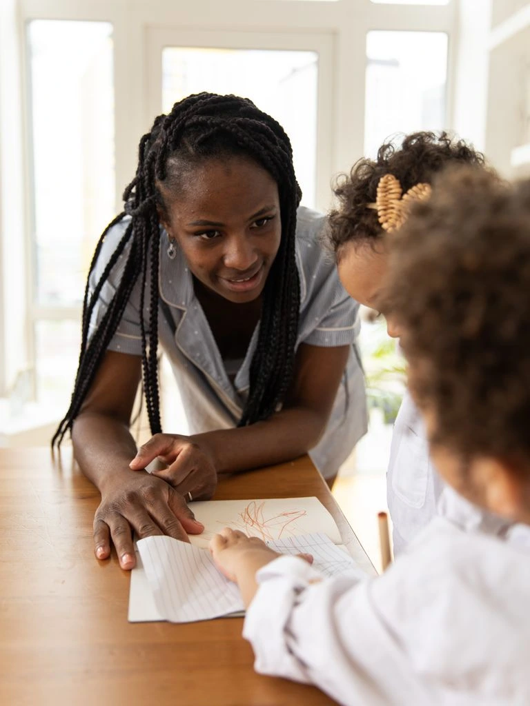 A homeschooling mother and her two children review classical textbooks at their kitchen table beside a packed moving box and a sealed transcript envelope.